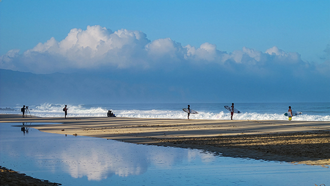 Three surfers line up to enter the waves on the North Shore of Oahu, while three sets of onlookers spectate.