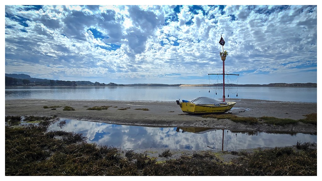 A view across the back bay from the direction of Los Osos toward the Morro Bay sand spit, including a small yellow sailboat beached on a sandbar in the foreground.