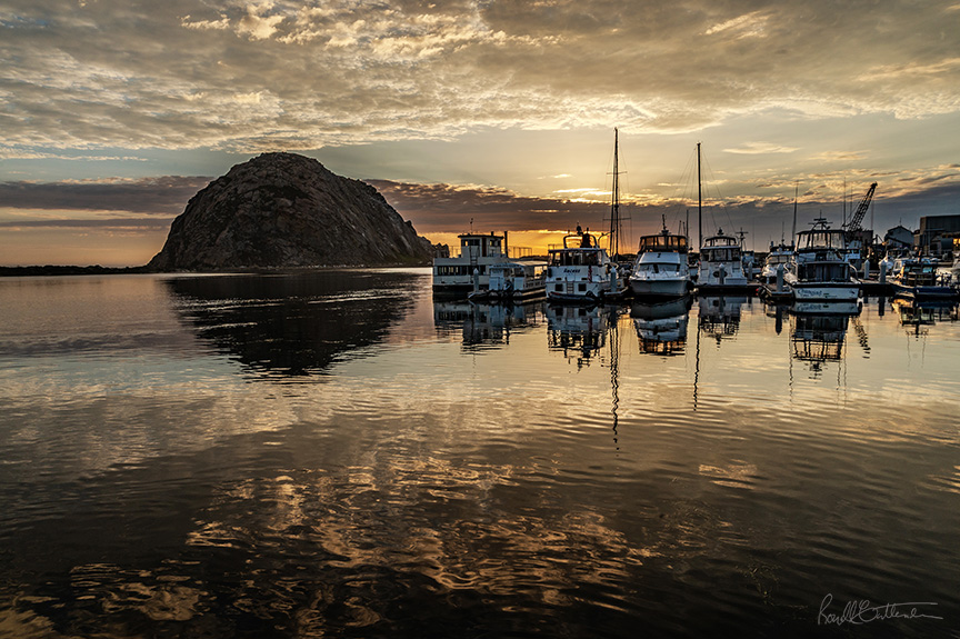 sunset over Morro Bay, with a line of boats in the middle distance and Morro Rock behind.