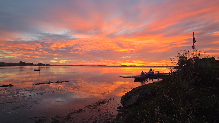 Sunset in hues of red, yellow, and blue paints the sky and waters of the Los Osos back bay, while a group of people look on from a pier in the right middle ground.
