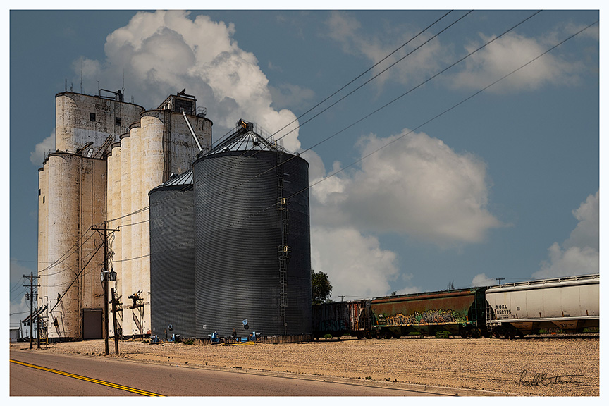 Commercial silos beside a train siding, with several cargo cars, against a backdrop of blue sky and clouds.