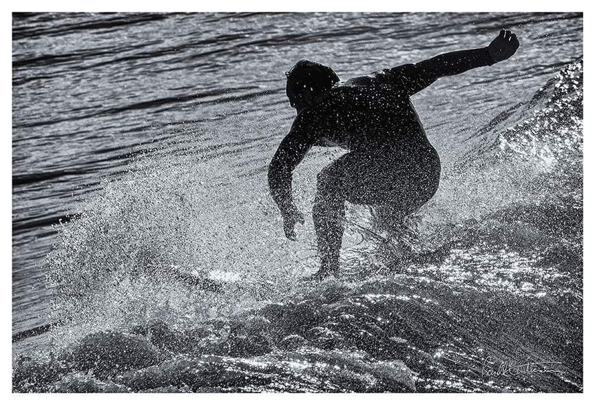 Striking a heroic pose, a surfer in silhouette finishes his run in a spray of water.